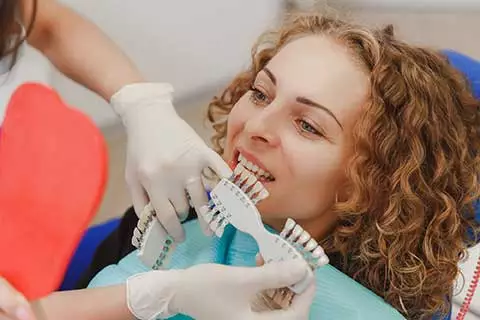 woman sits in a dental chair while the dentist compares which shade of tooth color matches the patient's teeth